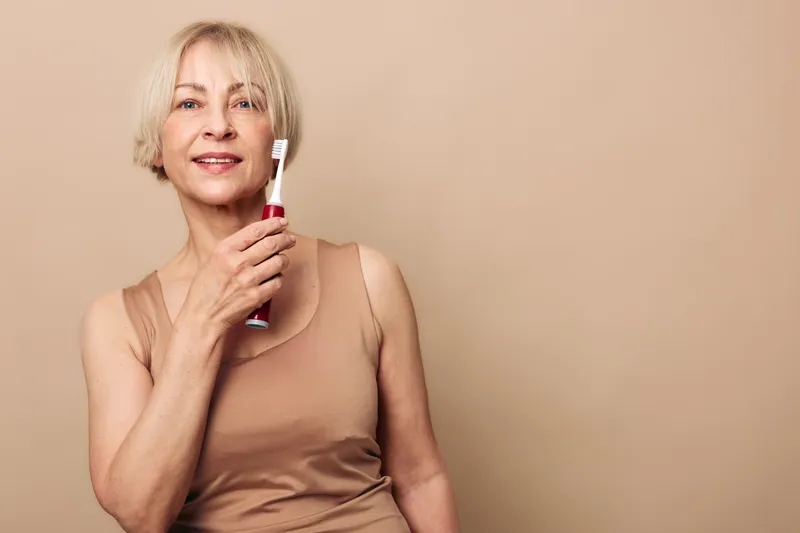 Mature woman with short blonde hair holding electric toothbrush near face, smiling gently. Neutral background and casual outfit emphasize dental hygiene and healthy lifestyle concept.