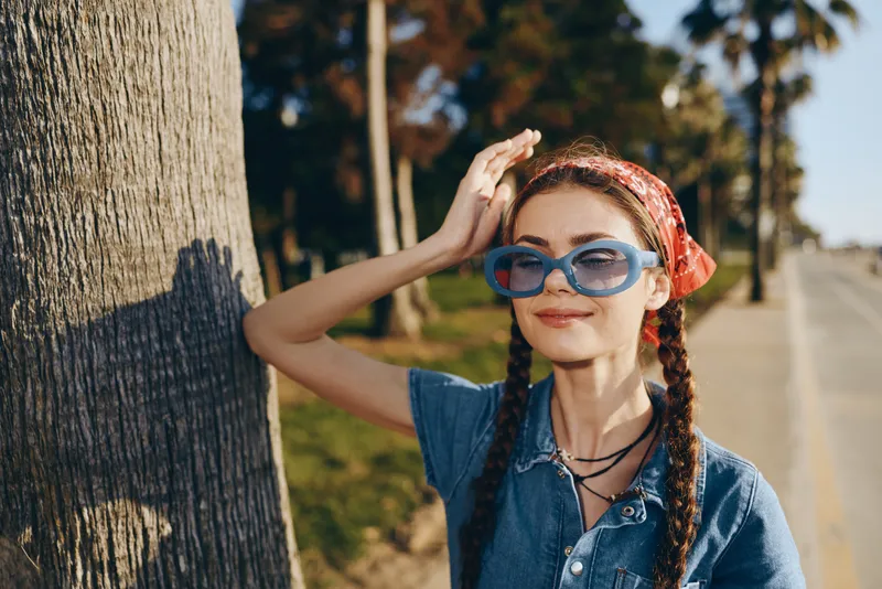 smiling woman wearing blue sunglasses and red headscarf standing near palm tree in sunny outdoor setting with casual denim shirt and braided hair
