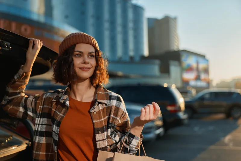 Woman shopping in city at golden hour, smiling by an open car trunk with bags and beanie, a candid lifestyle portrait conveying authenticity, mindful living and emotional storytelling.