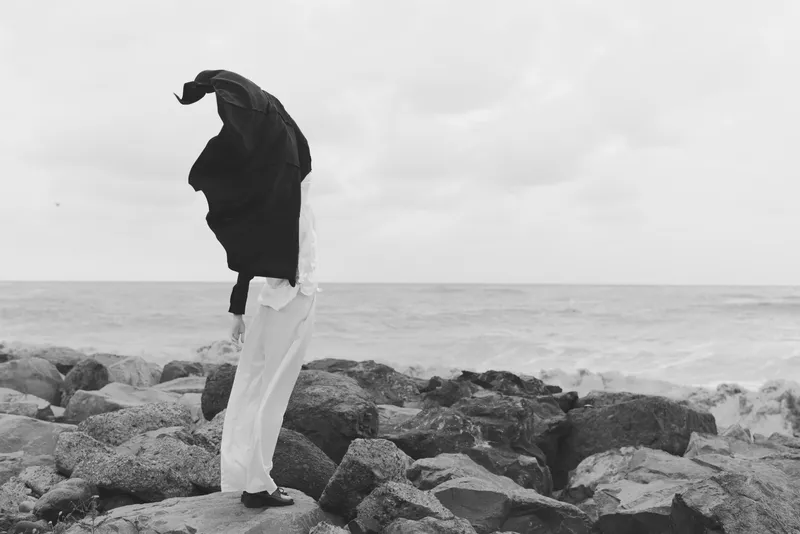 Monochrome scene of a hooded figure standing on jagged rocks by the sea, scarf flapping in the wind, capturing solitude, resilience and coastal mood against wide cloudy horizon