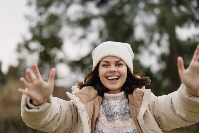 Woman in winter outerwear outdoors with outstretched arms, smiling joyfully, wearing a beanie and puffy jacket in a chilly outdoor setting for a friendly seasonal scene