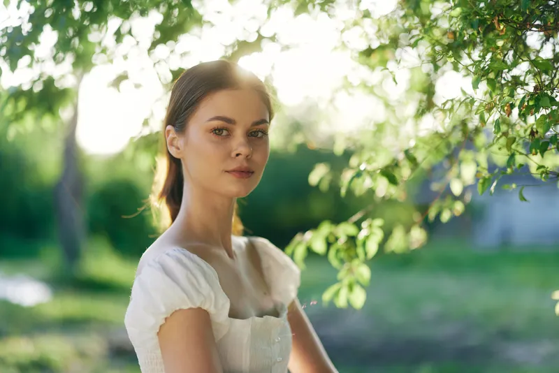 Young woman in a white dress with soft sunlight filtering through green leaves, capturing the essence of a warm summer day