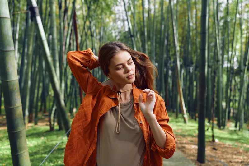 Woman portrait in a bamboo forest, nature and fashion blend as a serene outdoor model poses among green foliage and sunlight, wearing an orange jacket for a peaceful, meditative mood.