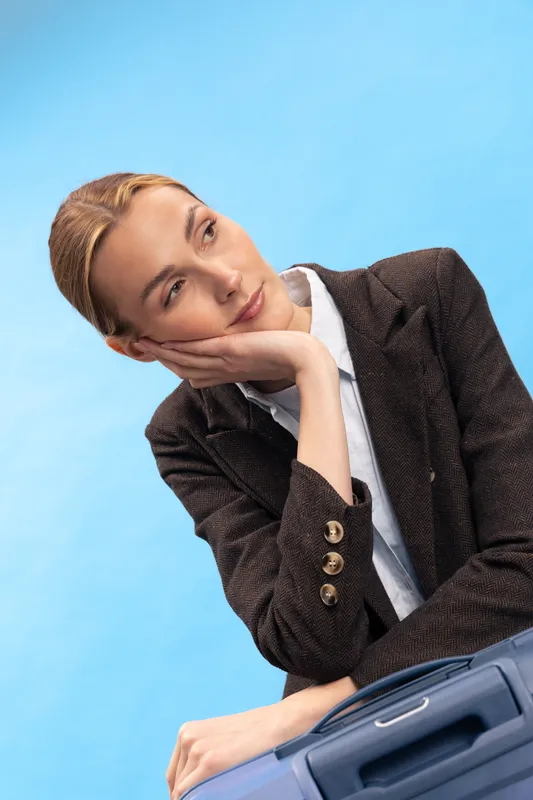 Thoughtful young woman in brown blazer rests her head on hand, sitting with suitcase on blue background. Concept of travel, waiting, and contemplation in casual fashion.