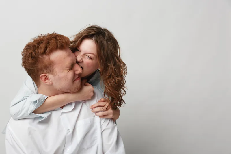 Playful young couple in white shirts share a joyful moment, woman hugging man from behind and pretending to bite his cheek. Concept of love, fun, affection, and relationship happiness.