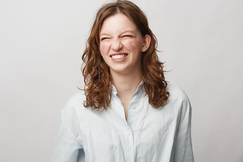 Young woman with curly hair smiles with closed eyes wearing white shirt on light background. Concept of happiness, natural beauty, casual style, and positive emotions.
