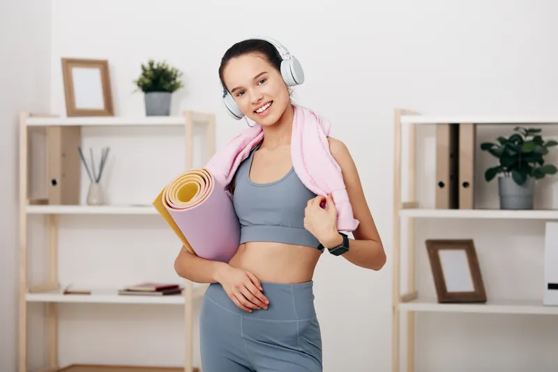 Young woman in a gym wearing gray sportswear, headphones, and a towel around her neck, holding a rolled mat ready for a workout during a fitness session.