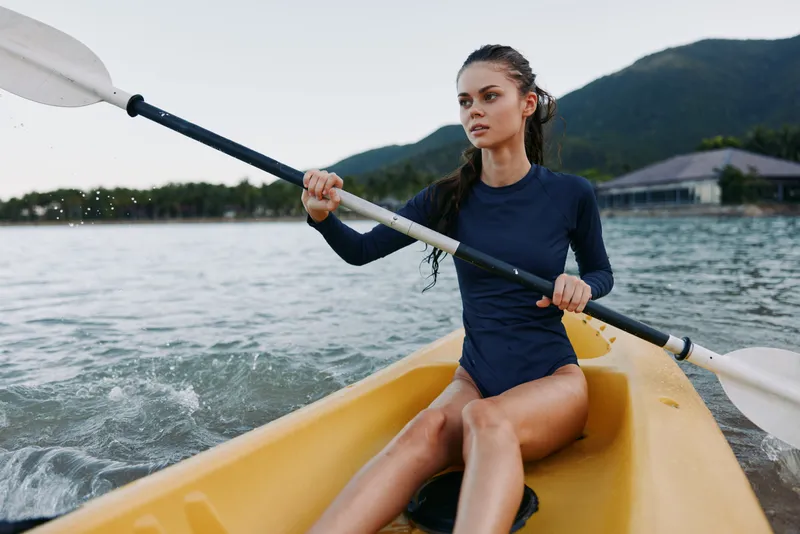 Focused female kayaker paddles a yellow kayak across calm water, gripping a white paddle, wearing a dark blue swimsuit with mountains in the background.