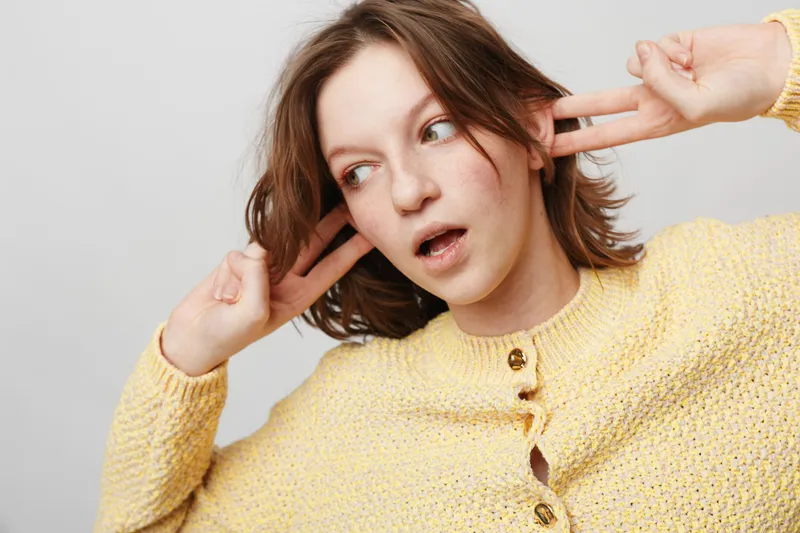 Young woman in yellow sweater covers ears with fingers and looks away with open mouth. Concept of annoyance, noise, discomfort, and emotional reaction in casual studio setting.