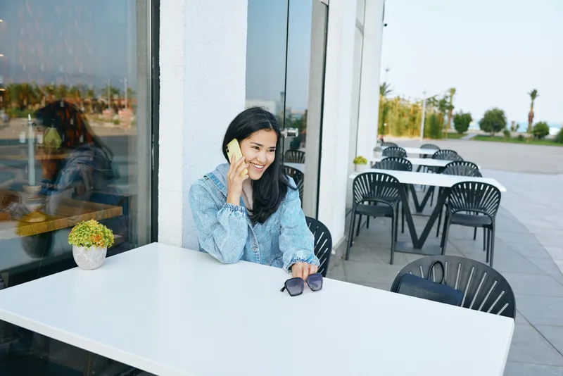 Woman smiling while talking on phone at an outdoor cafe, white table, sunglasses beside, sunny patio, glass storefront, casual urban vibe, relaxed daytime moment