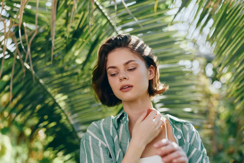 A woman in a green striped shirt stands among palm leaves as sunlight filters through, creating a calm, natural portrait with a relaxed expression.