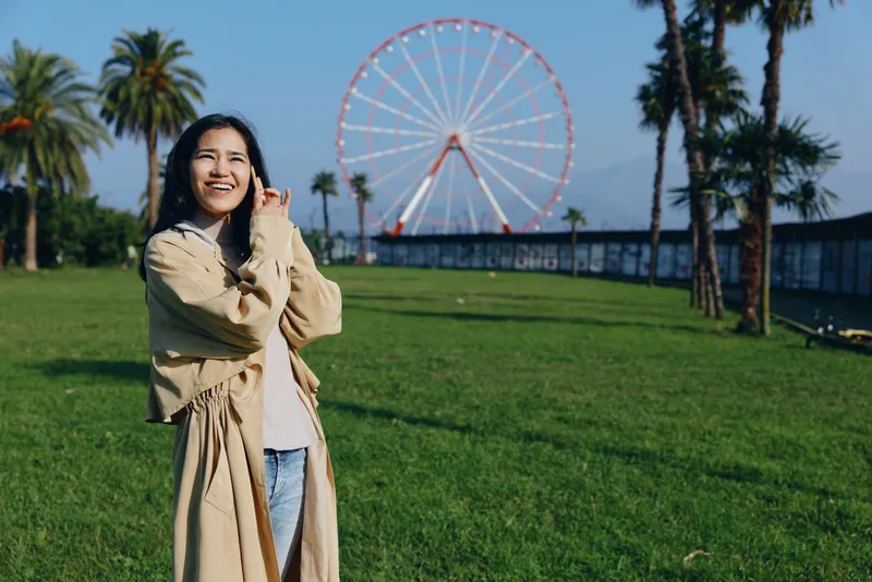 Woman wearing a beige trench coat smiles in a sunny park with a large ferris wheel in the background, capturing a joyful outdoor moment for lifestyle, travel, and leisure photography