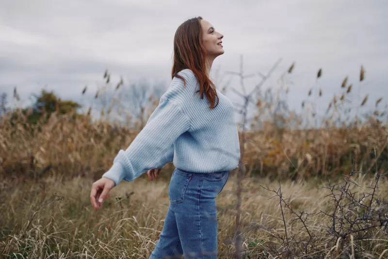 A solitary woman stands in windswept grass with arms outstretched, an authentic, genuine moment of calm and confidence captured in a realistic outdoor portrait conveying quiet assurance.