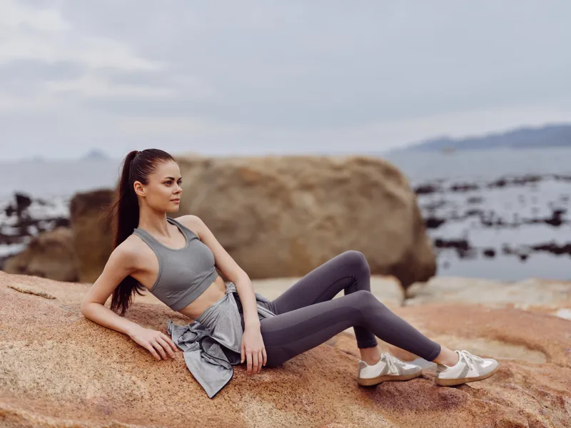 Young athletic woman sitting on rock by the seaside, wearing gray sportswear and sneakers, showcasing a relaxed and confident demeanor, ideal for fitness themes