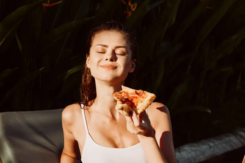 A relaxed woman enjoys a sunny outdoor moment, smiling with eyes closed as she eats pizza. A lifestyle portrait capturing leisure, warm light, and casual self care in a serene garden setting.