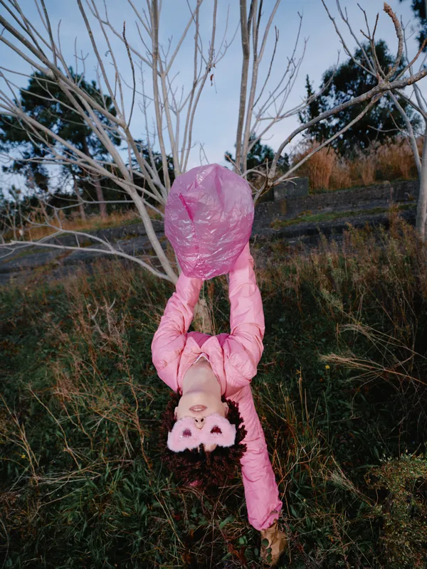 Little girl in a pink snowsuit hanging upside down from bare tree branches in a grassy field outdoors, playful adventurous portrait with soft natural light and woodland backdrop