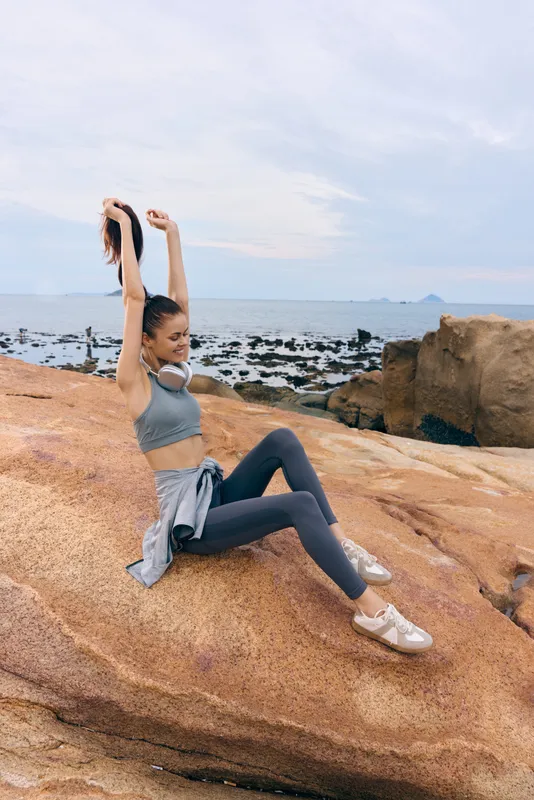 Young woman stretching in activewear on rocky beach during cloudy day, promoting fitness, well being, and outdoor lifestyle