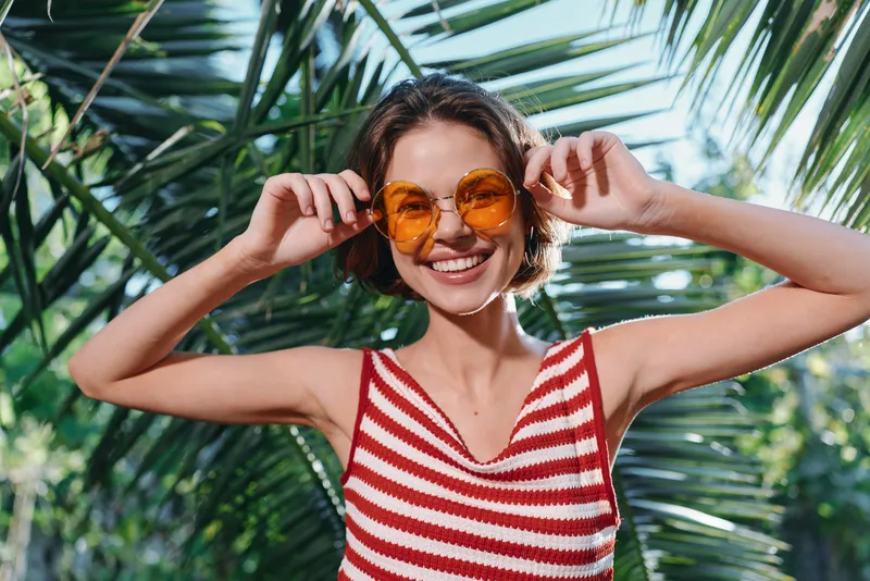 A cheerful woman wearing a striped red and white top stands outdoors beneath palm fronds. She smiles warmly, adjusts bright orange sunglasses, and exudes sunny summer energy.