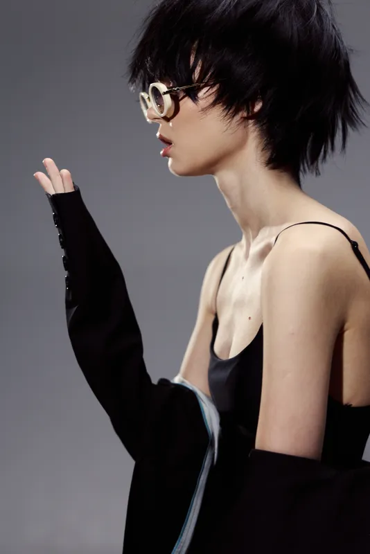 Stylish woman with short black hair wearing glasses and a black outfit in a minimalistic setting with neutral background.