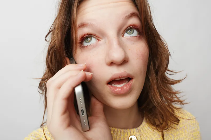 Young woman with red hair and freckles talks on smartphone, looking up with thoughtful expression. Casual lifestyle, communication, technology, and connection concept on white background.