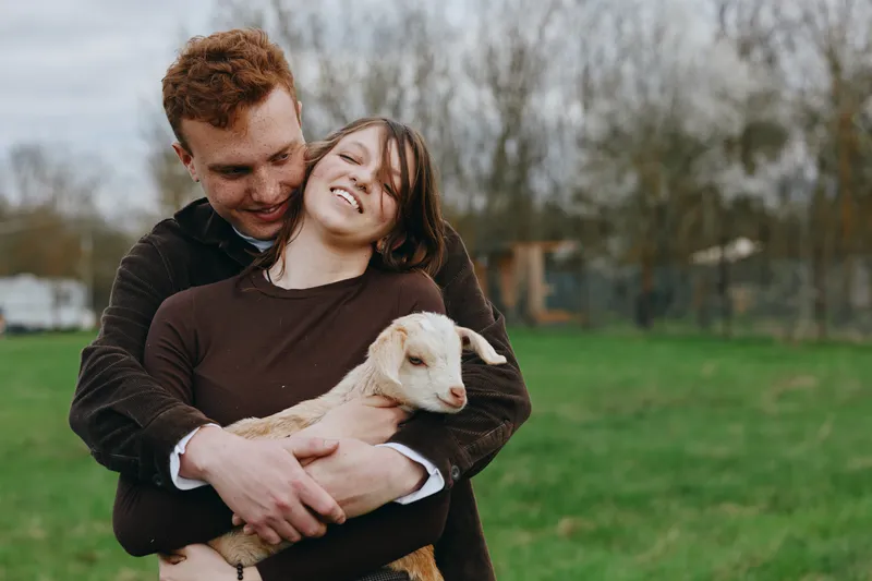 Young couple embraces outdoors while holding a small goat in their arms during a relaxed lifestyle moment in a green field on an overcast day.