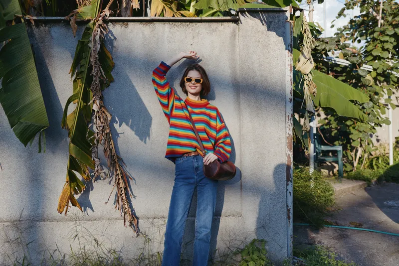 Woman in a colorful striped sweater and blue jeans wearing sunglasses, smiling against a concrete wall with tropical banana leaves, casual outfit and relaxed travel lifestyle vibe.