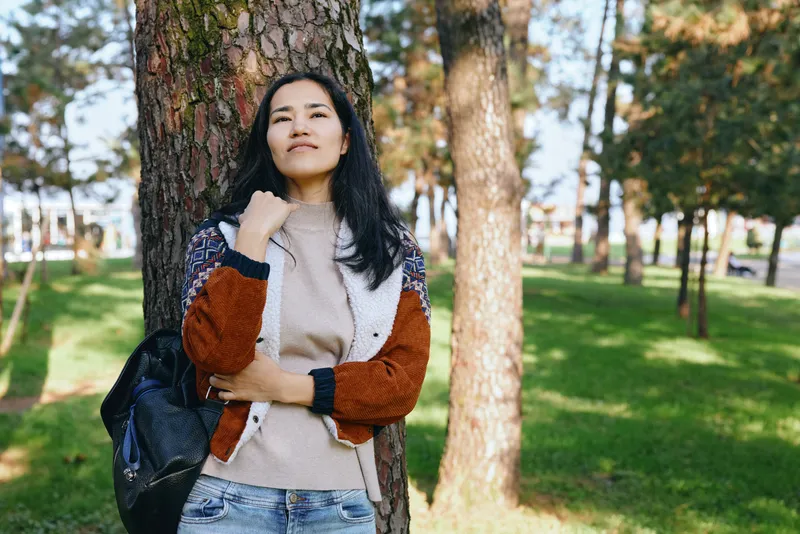 Woman in a cozy sweater and jacket stands outdoors in a park among tall trees, carrying a backpack, gazing upward with a relaxed, confident pose amid sunlight and gentle breeze in a calm, scenic