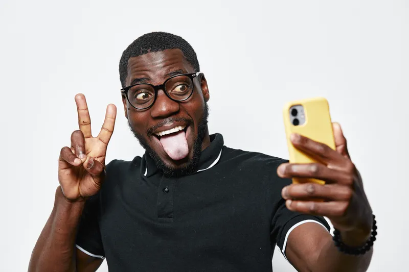 Black man with dark skin taking selfie with smartphone showing peace sign and sticking out tongue. Casual style, glasses, happy expression on white background.