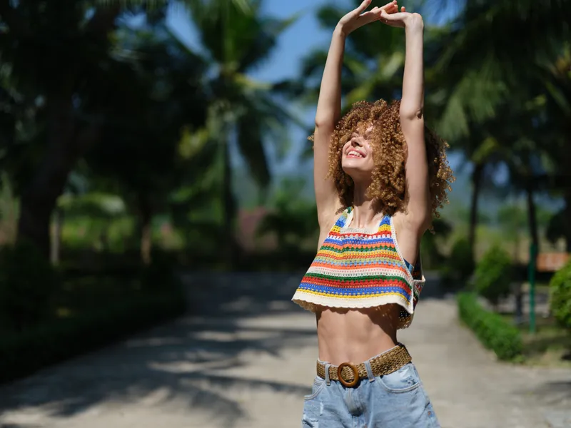 Smiling woman stretching outdoors in colorful summer clothing, enjoying sunshine with tropical greenery in the background, relaxed and happy.