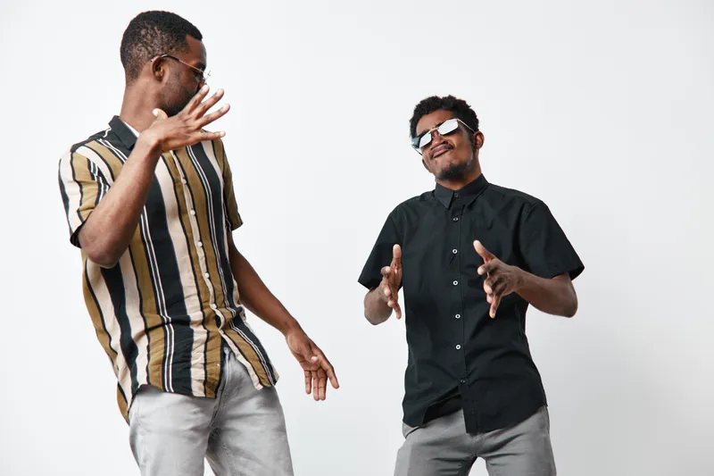 Two african men with dark skin dancing and having fun together in casual clothes and sunglasses on white background in studio. Friendship and joy concept.