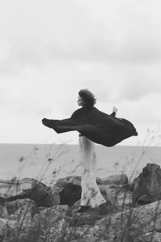 Cape billows as a figure stands on rocks beside the sea, wind swept landscape showing freedom, strength and calm in monochrome, capturing resolve