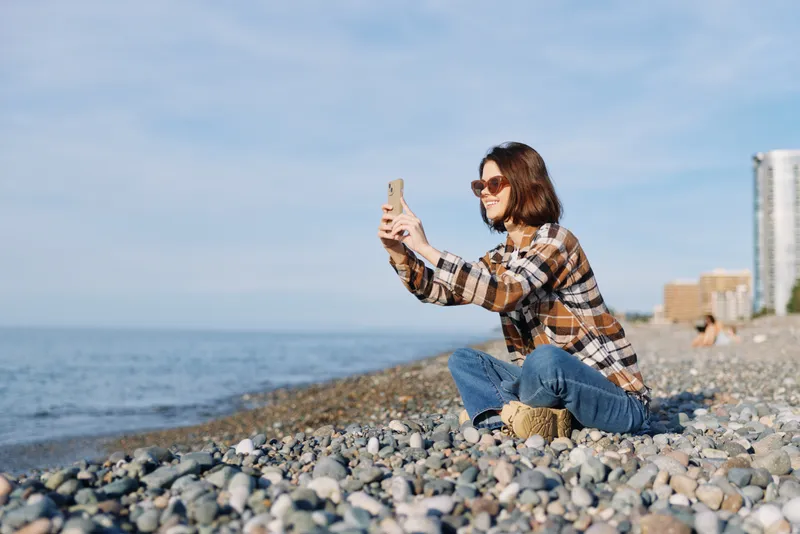 Person taking a selfie on a pebbled beach with a phone, wearing a casual plaid shirt and jeans; sunny outdoors with waves and distant buildings; relaxed moment captured for travel, lifestyle, and
