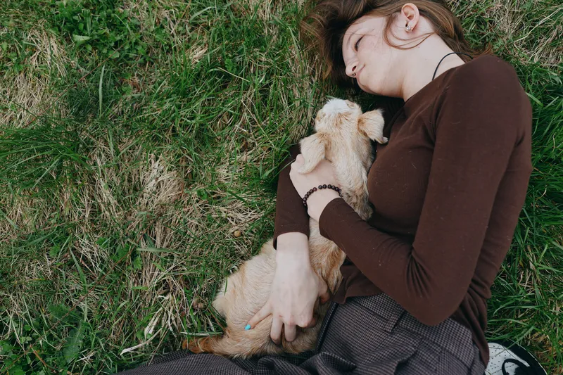 Woman lying on grass, cuddling a dog, enjoying a peaceful outdoor moment, relaxed expression, casual clothing, natural light, intimate companionship, slow living, comforting pet presence