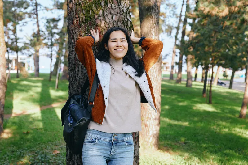 Woman leaning against a tree in a sunlit park, smiling in a casual autumn outfit, relaxed outdoor moment, cheerful and carefree.