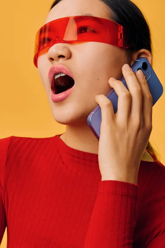 Woman wearing red glasses and red shirt holding smartphone near ear with open mouth speaking on yellow background. Female person in casual clothing making a call with expression.