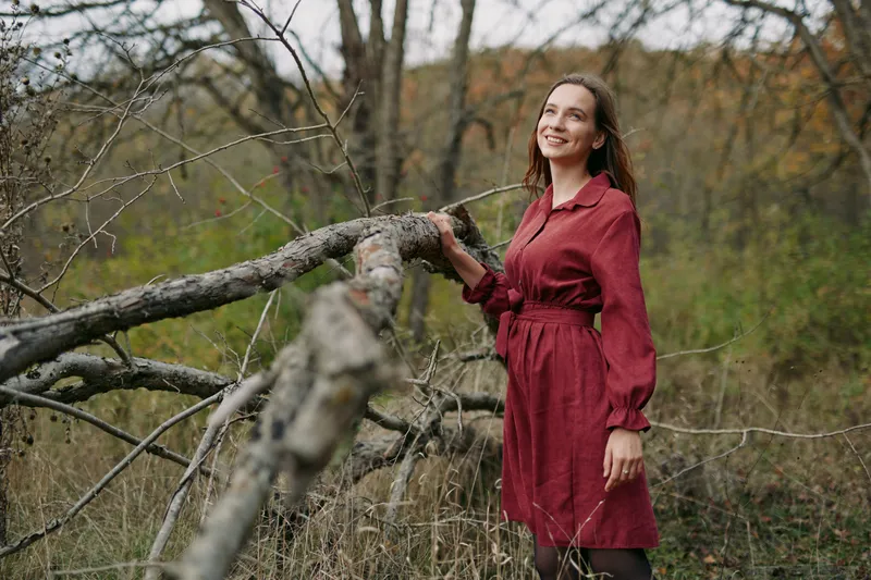 A woman in a burgundy dress stands beside a weathered fallen tree, conveying authenticity and quiet confidence in a natural outdoor setting.
