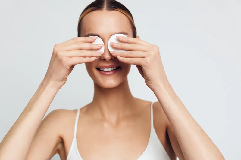 Smiling woman with clean skin holding cotton pads over her eyes on white background. Concept of skincare, facial cleansing, and beauty routine. Soft natural lighting.