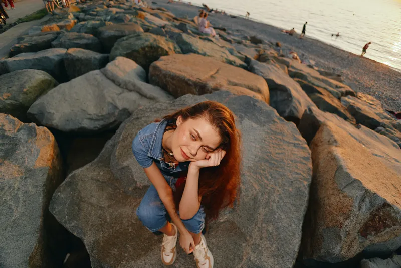 lifestyle woman in modern boho-western denim street style sitting relaxed on large beach rocks with warm film color tones during sunset