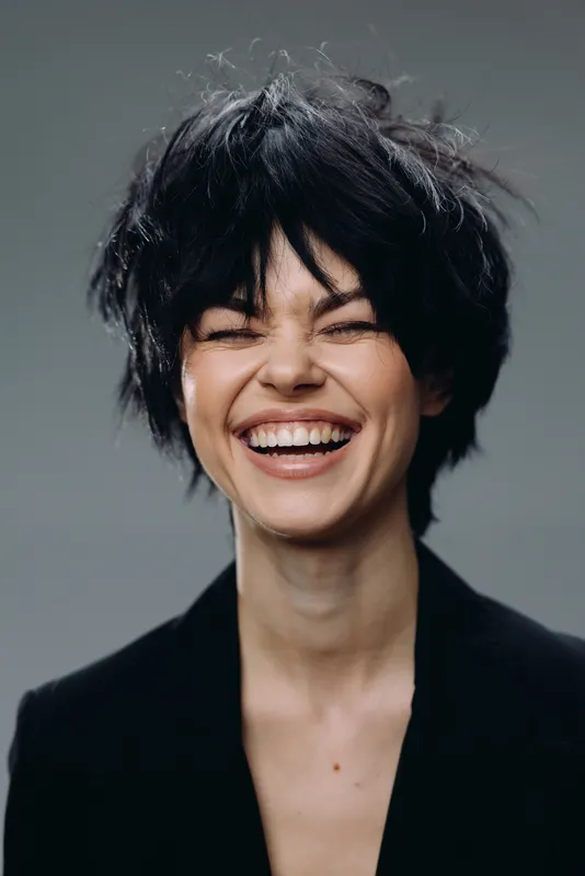 smiling woman with short black hair laughing joyfully in studio, casual black jacket, energetic and happy expression