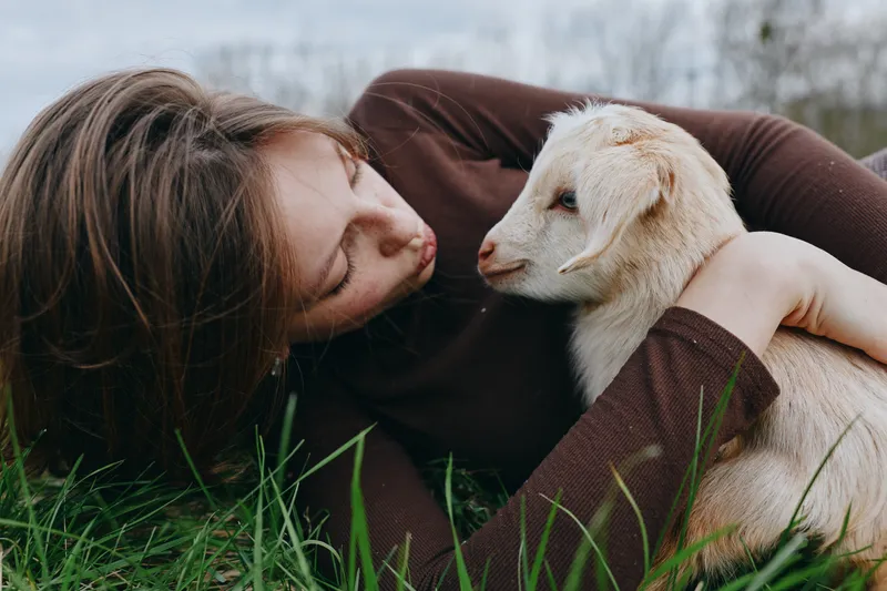 A woman lies in lush grass, embracing a curious goat kid in a peaceful outdoor setting. Warm natural light highlights affection, companionship, and lifestyle, blending nature, care, and simple