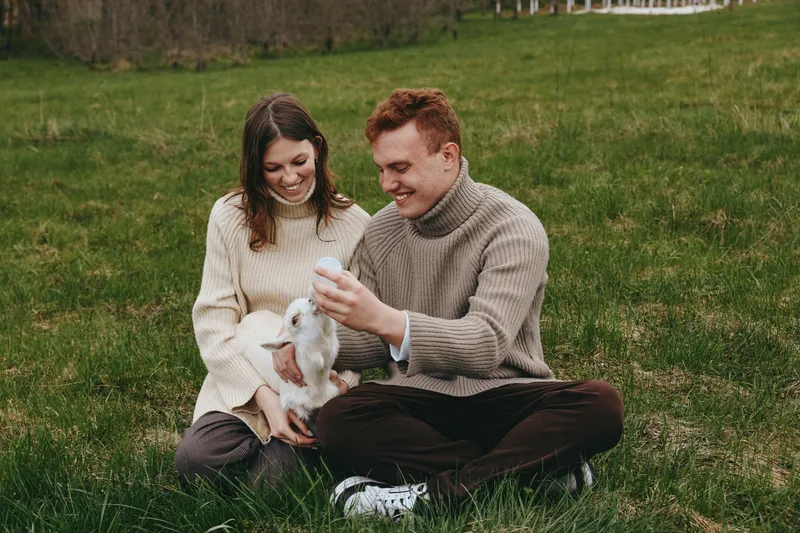 A joyful couple sits on grass with a young goat, feeding and bonding in an open field. A warm lifestyle moment highlighting care, connection, and outdoor life with animals.