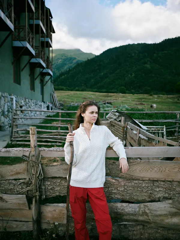 Woman wearing a white sweater and red pants stands beside a rustic fence on a countryside farm, with green pastures and mountains in the distance, a calm outdoor lifestyle scene