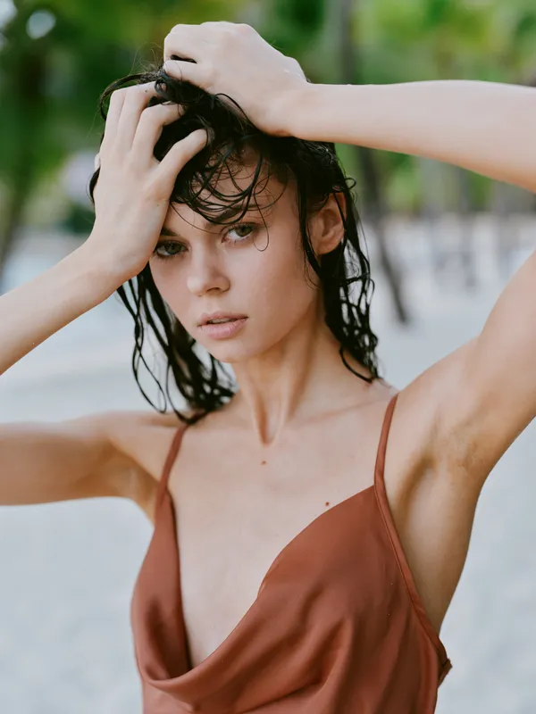 Close portrait of a woman with wet hair, standing on a sunlit beach in a rust top. Intense gaze and natural vibe capture bold confidence and summer mood.
