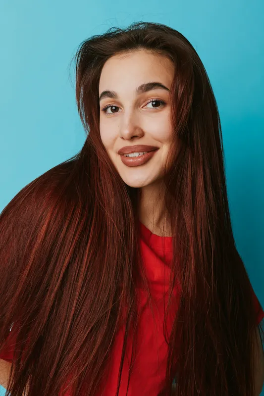 Young woman with long straight hair and natural makeup smiling against blue background. Portrait of happy female in red shirt looking at camera with confident expression and relaxed pose.