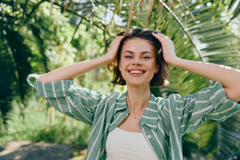 Smiling young woman outdoors adjusting hair, natural light, green foliage background, casual striped shirt, relaxed mood, candid portrait.