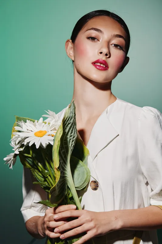Young stylish woman with clear skin and bold red lips holds fresh white daisies. Soft lighting highlights fashion makeup and creative beauty concept on pale green background.