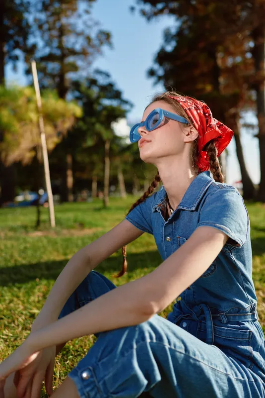 lifestyle woman in modern boho-western denim street style wearing red bandana and blue sunglasses, enjoying sunny outdoor park in warm natural light with film color effect