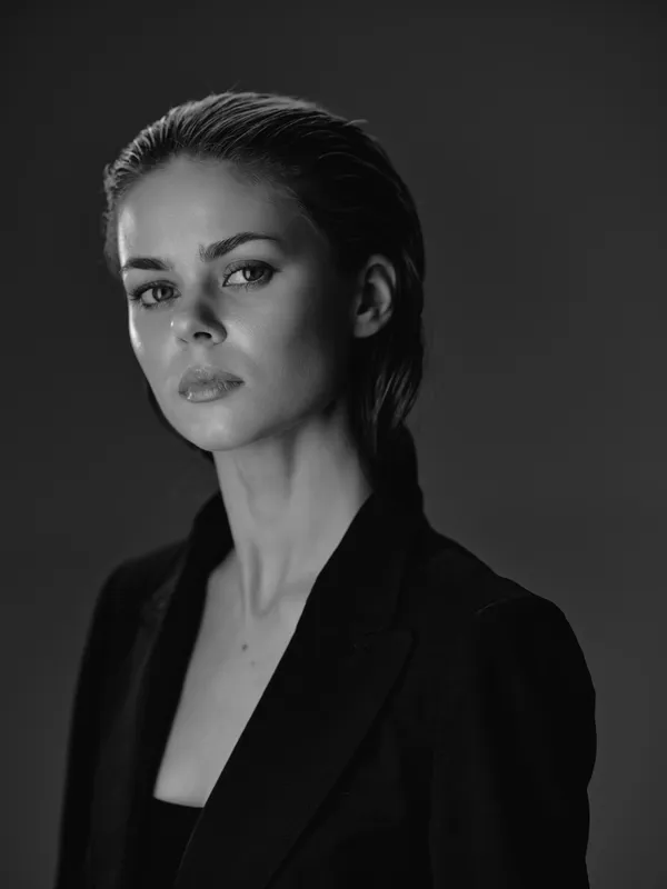 Black and white portrait of a serious woman in a blazer with slicked-back hair and neutral expression in studio lighting.