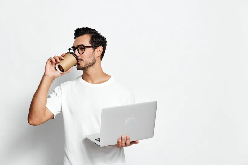 Young man in casual white t-shirt holds laptop and coffee cup, looking thoughtfully at the side. Modern lifestyle concept with minimalistic background.