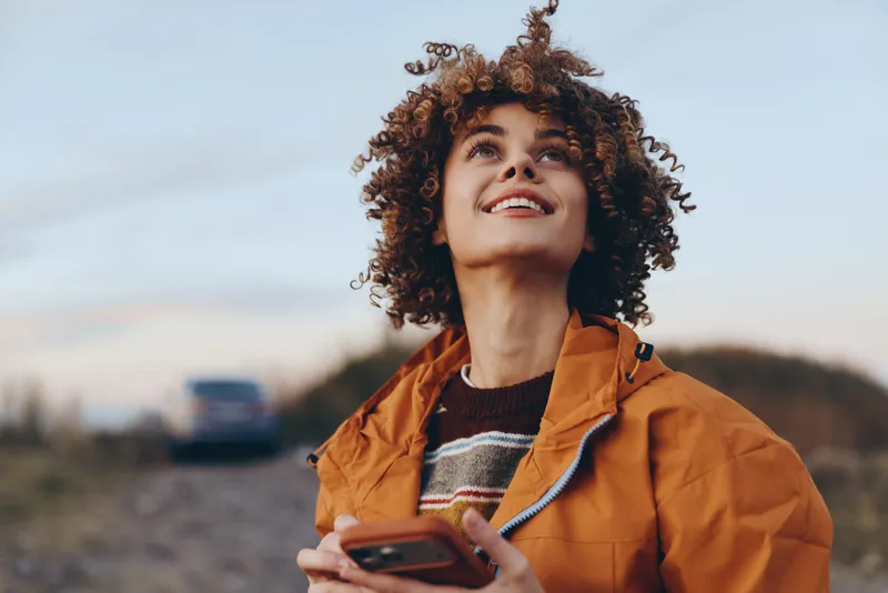 Woman outdoors, curly hair, smartphone in hand, orange jacket, lifestyle smile at warm golden sunset in nature, relaxed posture, soft wind, candid portrait, distant hills blurred